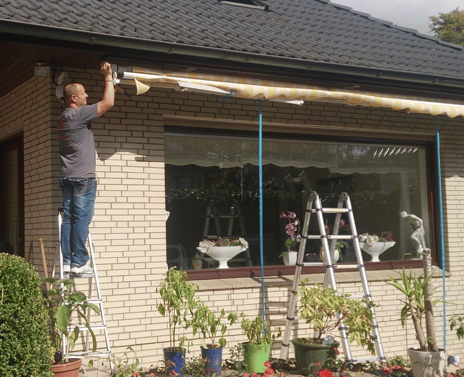 Mann steht auf einer Leiter und installiert eine Markise über einem Fenster eines Wohnhauses.